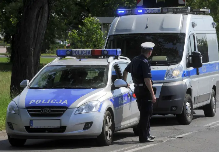 Policjant stoi obok radiowozu i furgonetki policyjnej z włączonymi sygnałami świetlnymi.