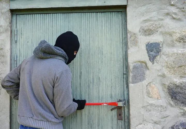 Osoba w masce próbuje wyważyć drzwi łomem. To klasyczny przykład kradzieży z włamaniem.