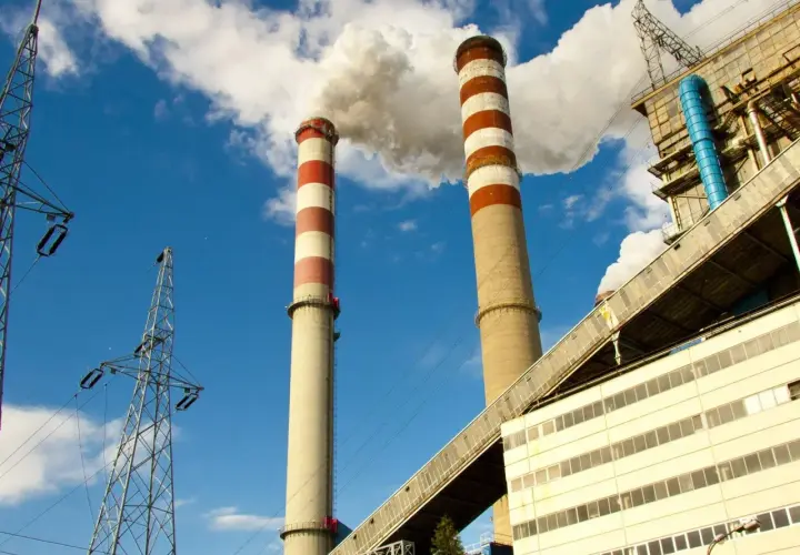 Dwie wysokie, prążkowane kominy elektrowni emitują dym na tle błękitnego nieba. Obok widać linie energetyczne i budynki przemysłowe, tworzące obraz kluczowej infrastruktury.