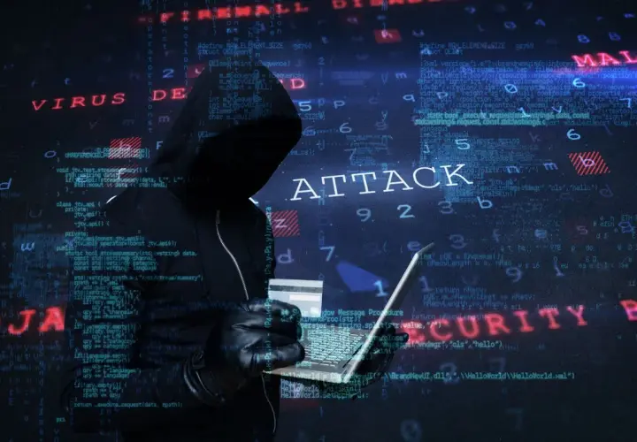 Sylwetka hakera w kapturze, trzymająca laptopa i kartę kredytową. Na tle widać kod i napisy "ATTACK", "VIRUS", "MALWARE", "SECURITY", sugerujące atak hakerski.