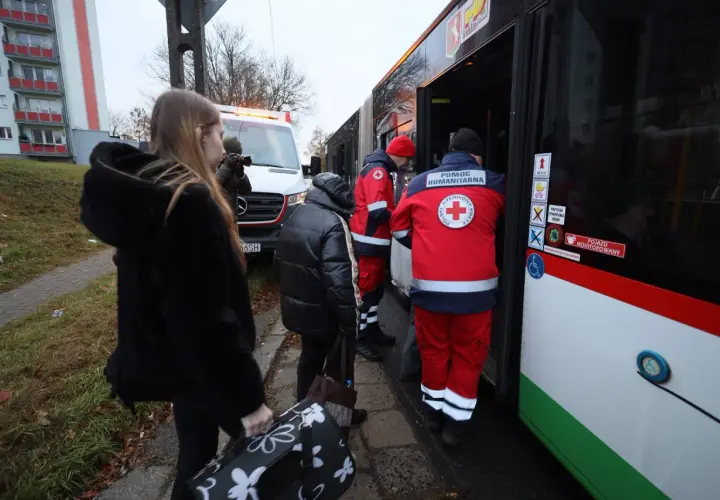 Osoby z bagażami wsiadają do autobusu z pomocą humanitarną. W tle biała furgonetka, przypominająca pojazd z lotniska, gdzie mógł wystąpić alarm.