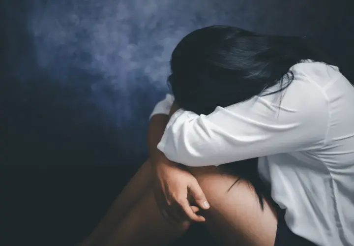Kobieta w białej koszuli skulona, przeżywająca traumę po gwałcie. Ciemne tło podkreśla jej cierpienie.