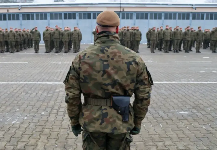 Żołnierz w mundurze moro i beretcie stoi na placu apelowym przed szeregiem innych żołnierzy.