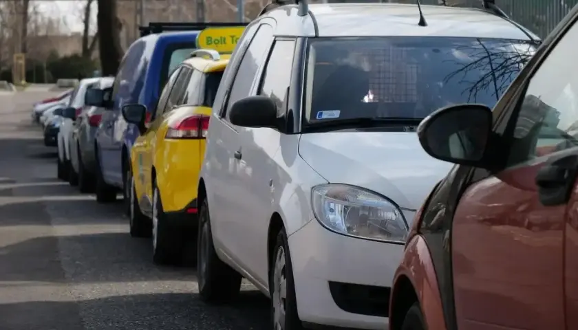 Rząd samochodów, w tym żółty Bolt Taxi, zaparkowany na chodniku.