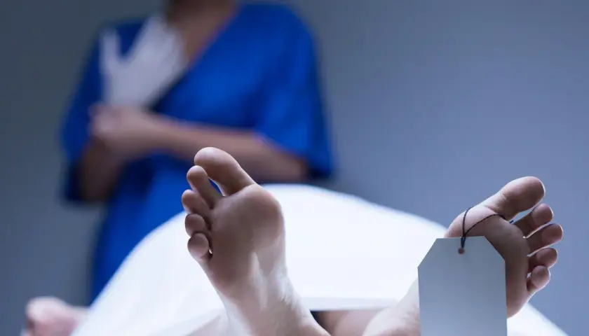 Stopy z metką identyfikacyjną, w tle osoba w niebieskiej bluzie zakłada rękawiczki. Widoczne plamy opadowe na skórze.