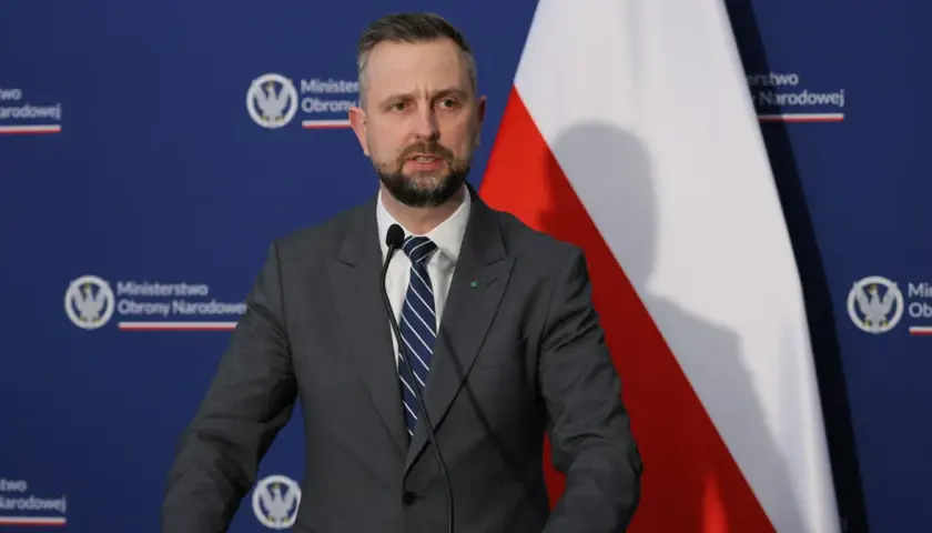 Mężczyzna w garniturze, minister obrony narodowej, przemawia przy mikrofonie na tle flagi Polski i logo ministerstwa.