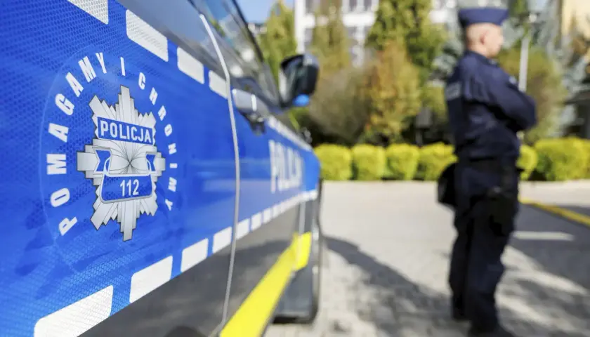 Policjant w mundurze stoi obok radiowozu z napisem "POLICJA" i numerem alarmowym 112.