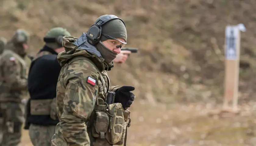 Żołnierz w mundurze maskującym, słuchawkach i okularach ochronnych celuje z pistoletu na strzelnicy.