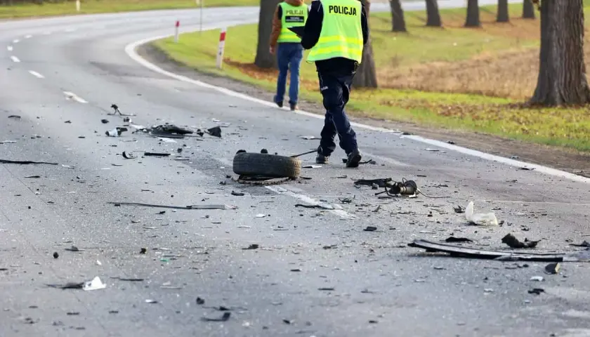 Na drodze po kolizji leżą szczątki, opona i elementy pojazdów. Policjanci w kamizelkach odblaskowych badają miejsce zdarzenia, być może szukając śladów ucieczki z miejsca kolizji.