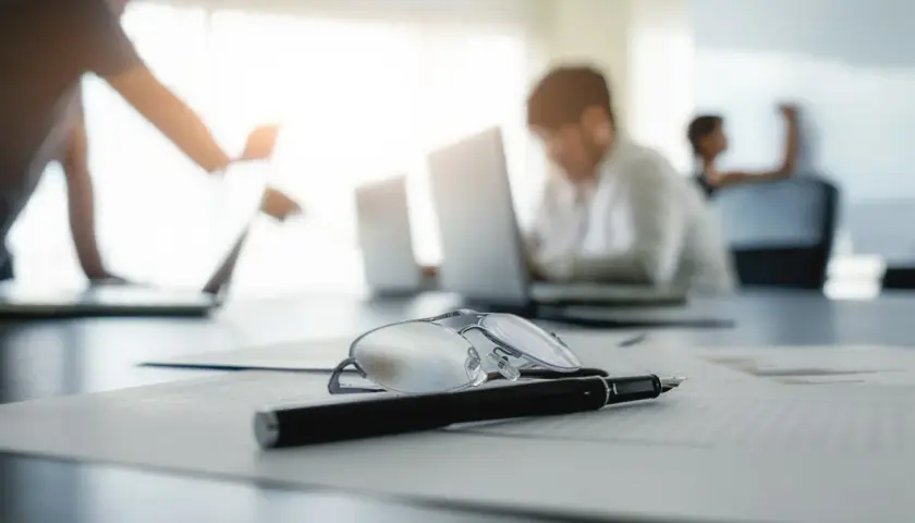 Okulary i długopis na dokumentach. W tle ludzie pracujący przy laptopach, być może praca na czarno.