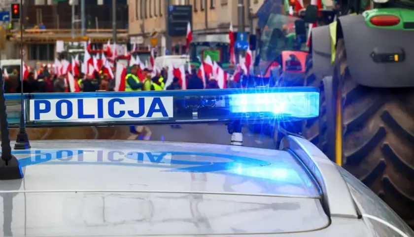 Policyjny radiowóz z włączonym sygnałem świetlnym na tle protestu rolników z flagami. Czy to jego jaka odprawa po 20 latach w policji?