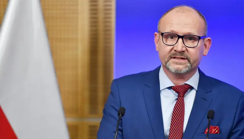 Prokurator krajowy w niebieskim garniturze, z okularami i czerwoną poszetką, przemawia przy mikrofonie. Obok widoczna flaga Polski.