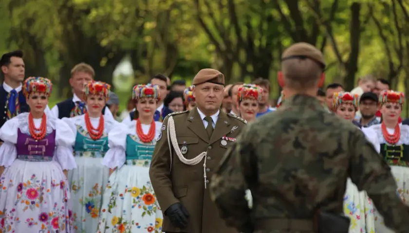 Żołnierz w brązowym capstrzyku i mundurze stoi przed grupą kobiet w tradycyjnych, kolorowych strojach ludowych.
