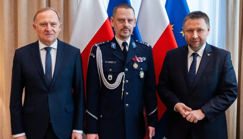 Trzech mężczyzn w garniturach i mundurze policyjnym stoi przed flagami Polski i UE.