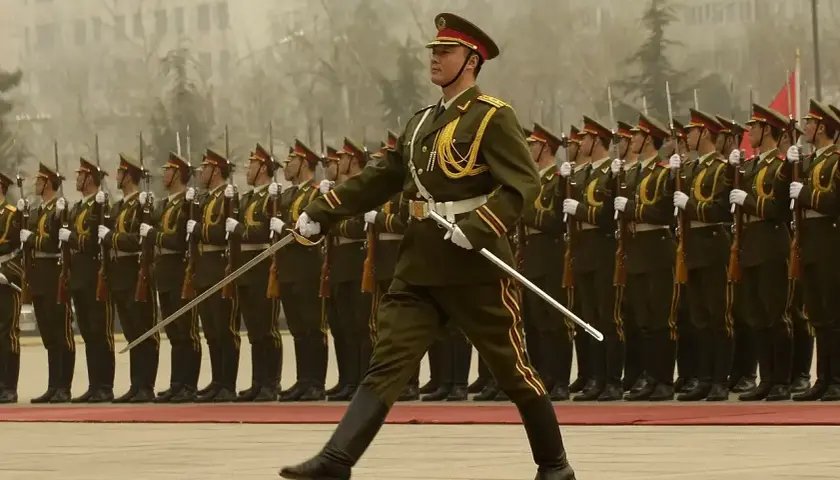 Kadet w galowym mundurze prowadzi defiladę, trzymając szablę. Za nim szeregi żołnierzy z karabinami.