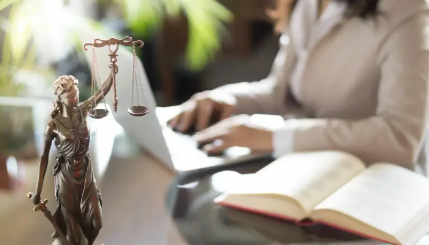 Figurka Temidy z wagą i mieczem, obok otwarta księga i osoba pracująca przy laptopie. Symbol sprawiedliwości i abolicji.