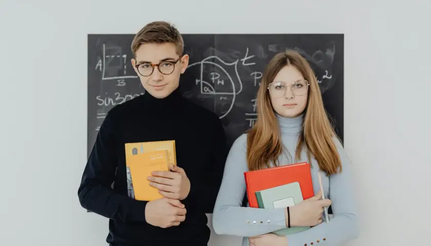 Młodociany uczeń i uczennica z książkami przed tablicą z matematycznymi wzorami.
