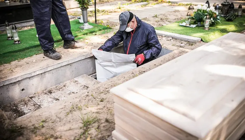 Pracownicy cmentarza przeprowadzają ekshumację, wyciągając biały worek z grobu. Obok leży drewniana trumna.