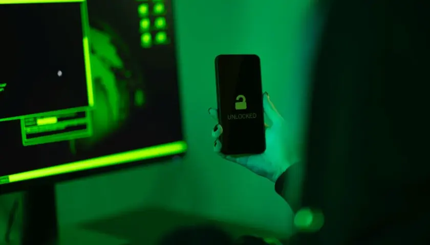Ręka trzyma odblokowany telefon na tle monitora z zielonym kodem. Jak zrobić, by policja nie namierzyła telefonu?