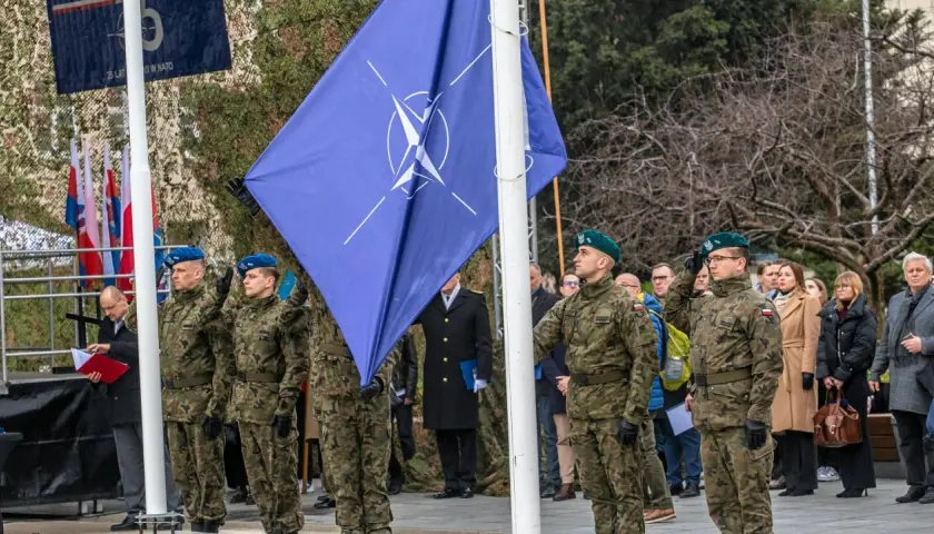 Uroczystość z flagą Organizacji Traktatu Północnoatlantyckiego. Żołnierze w mundurach oddają honory.