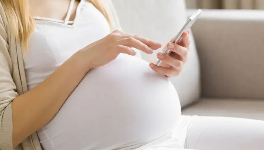Kobieta w 34. tygodniu ciąży przegląda coś na telefonie, siedząc na kanapie.