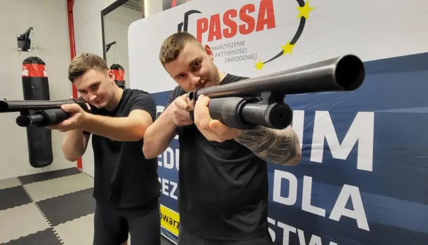 Dwóch mężczyzn ćwiczy strzelanie z broni gładkolufowej. Jeden z nich, kwalifikowany pracownik ochrony, celuje w obiektyw.
