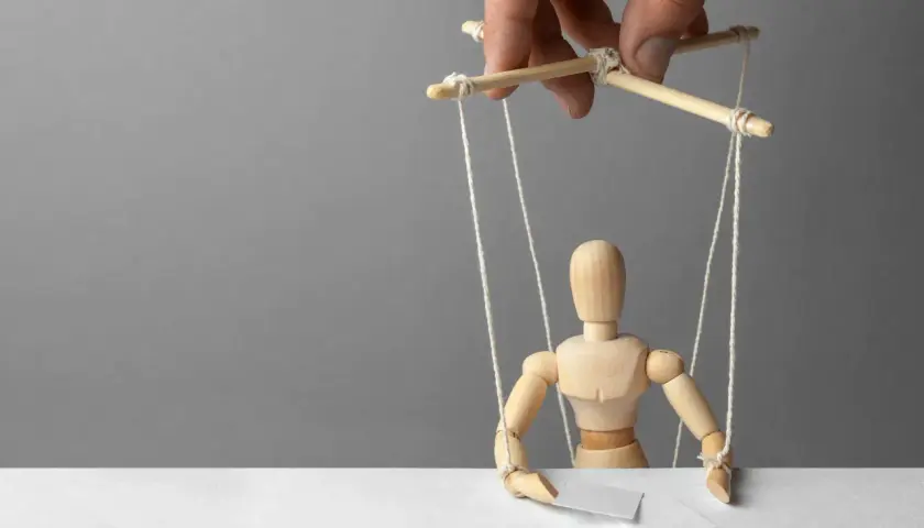 Drewniany ludzik-marionetka, sterowany przez ludzką dłoń, symbolizuje podżeganie i manipulację.