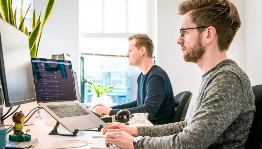 Dwóch programistów pracuje przy komputerach. Jeden z nich ma okulary, co może sugerować refundację okularów przez pracodawcę.