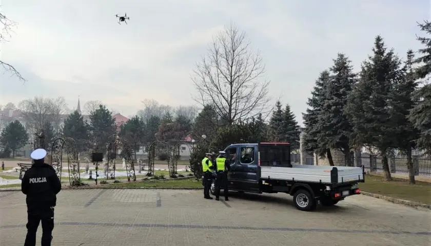 Dron policyjny w powietrzu, obok radiowozu i policjantów.
