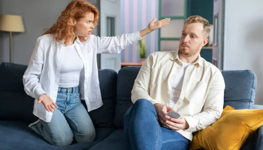 Kobieta z burzą rudych włosów, w białej koszuli i jeansach, gestykuluje, wyrażając swoje emocje. Mężczyzna obok, w jasnej koszuli, patrzy w bok, trzymając telefon. Scena przypomina sytuację z art. 193 kk.