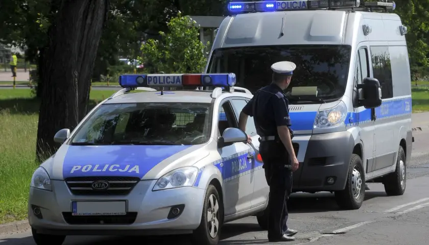 Policjant stoi obok radiowozu i furgonetki policyjnej z włączonymi sygnałami świetlnymi.