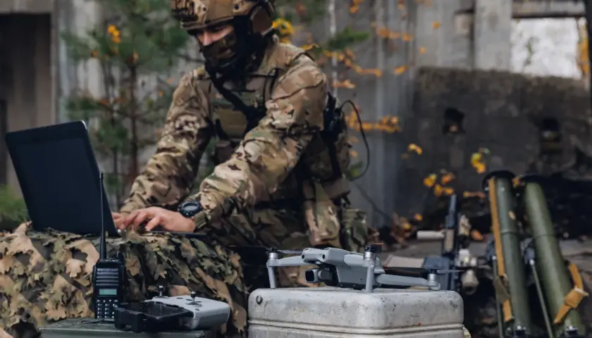 Żołnierz w kamuflażu pracuje na laptopie podczas ćwiczeń wojskowych. Obok leży dron i radiostacja.