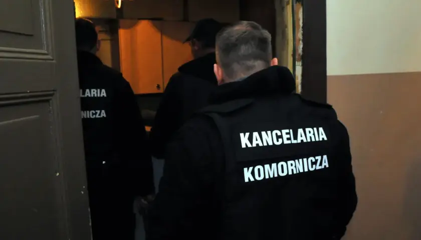 Trzech mężczyzn w czarnych kamizelkach z napisem "KANCELARIA KOMORNICZA" wchodzi do pomieszczenia. Prawdopodobnie to postępowanie egzekucyjne.