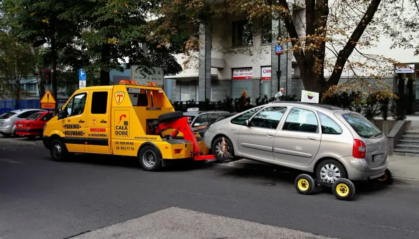Żółty holownik zabiera srebrny samochód, który zapewne otrzymał mandat za parkowanie.