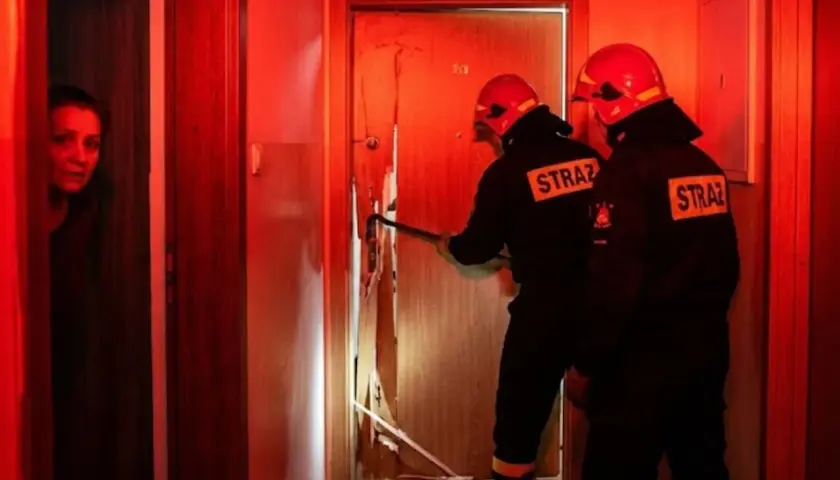 Strażacy wyważają drzwi, gdy kobieta patrzy z niepokojem. Sytuacja, kiedy policja może wyważyć drzwi, by ratować życie lub zapobiec przestępstwu.