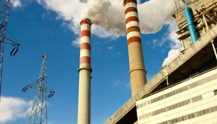 Dwie wysokie, prążkowane kominy elektrowni emitują dym na tle błękitnego nieba. Obok widać linie energetyczne i budynki przemysłowe, tworzące obraz kluczowej infrastruktury.