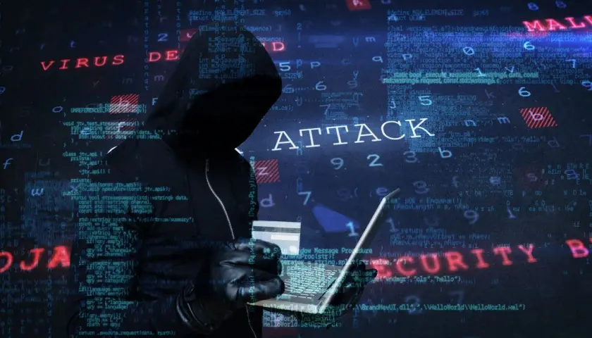 Sylwetka hakera w kapturze, trzymająca laptopa i kartę kredytową. Na tle widać kod i napisy "ATTACK", "VIRUS", "MALWARE", "SECURITY", sugerujące atak hakerski.