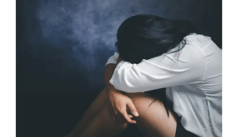 Kobieta w białej koszuli skulona, przeżywająca traumę po gwałcie. Ciemne tło podkreśla jej cierpienie.