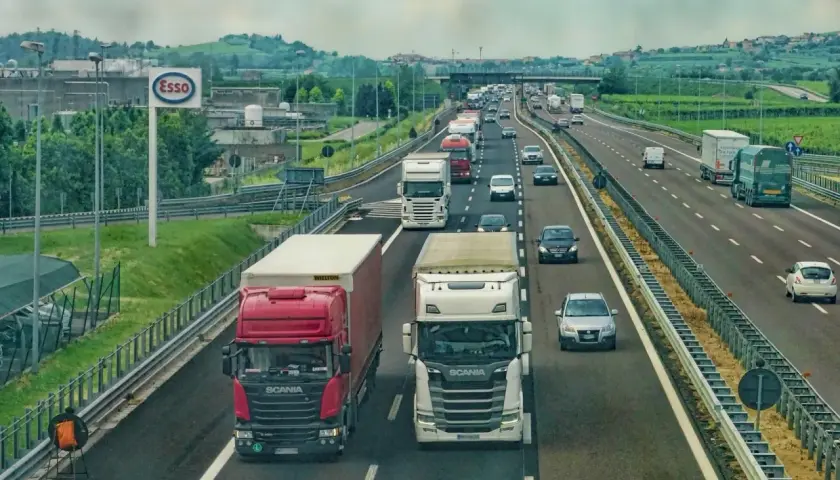 Ciężarówki na autostradzie, jedne wymijają, inne omijają. W tle znak Esso i zielone wzgórza.