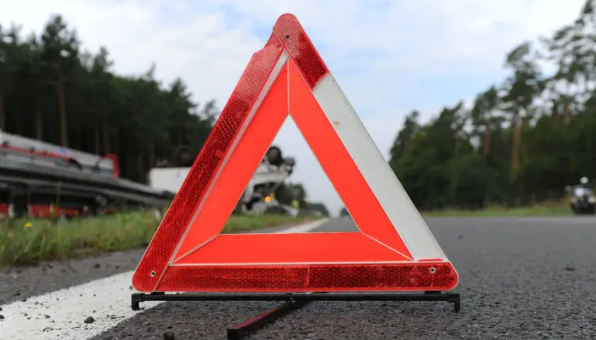 Trójkąt ostrzegawczy, część obowiązkowego wyposażenia samochodu, stoi na poboczu drogi. W tle widać przewrócony samochód ciężarowy.