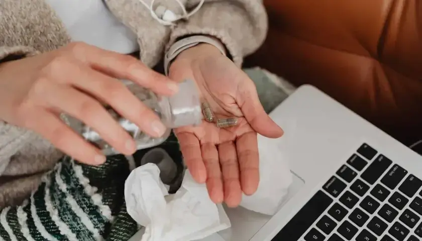 Dłonie z kapsułkami leków, obok laptop i chusteczki. Kto płaci za L4, gdy choroba dopada w domu?