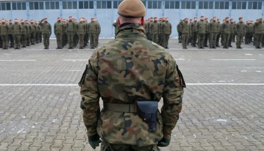 Żołnierz w mundurze moro i beretcie stoi na placu apelowym przed szeregiem innych żołnierzy.