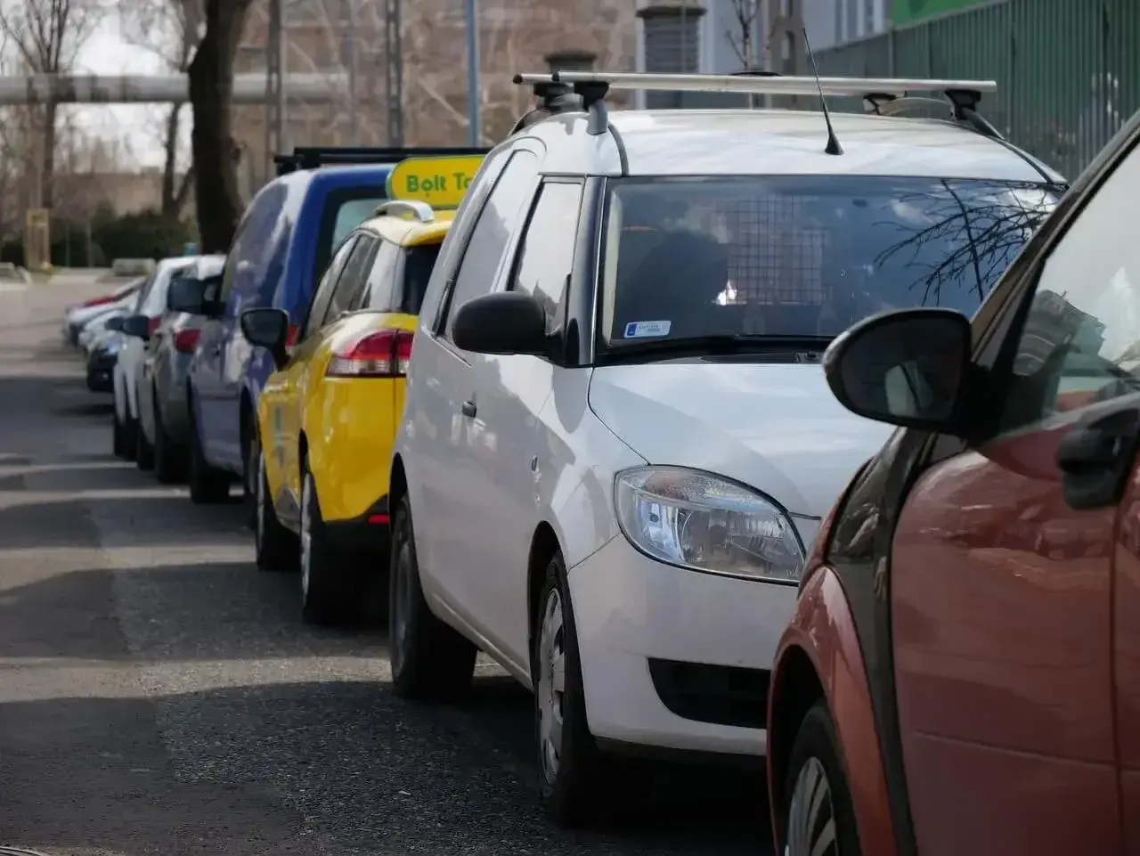Rząd samochodów, w tym żółty Bolt Taxi, zaparkowany na chodniku.