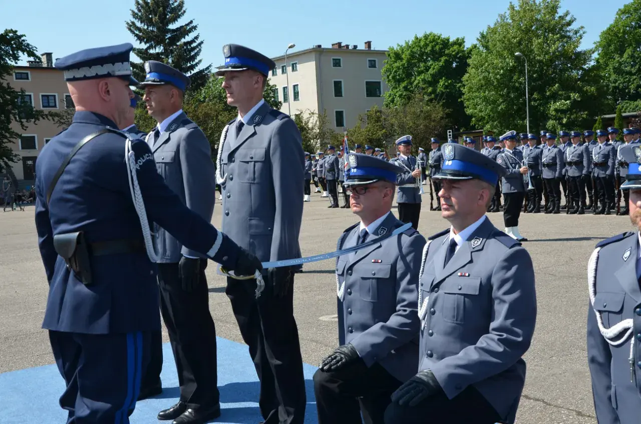 Uroczyste mianowanie na nowych zasadach. Policjanci w galowych mundurach, jeden klęczy, otrzymując szablę.