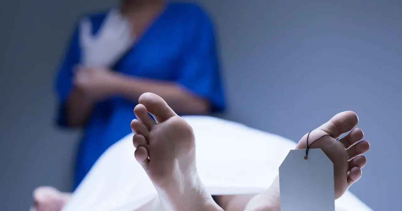 Stopy z metką identyfikacyjną, w tle osoba w niebieskiej bluzie zakłada rękawiczki. Widoczne plamy opadowe na skórze.