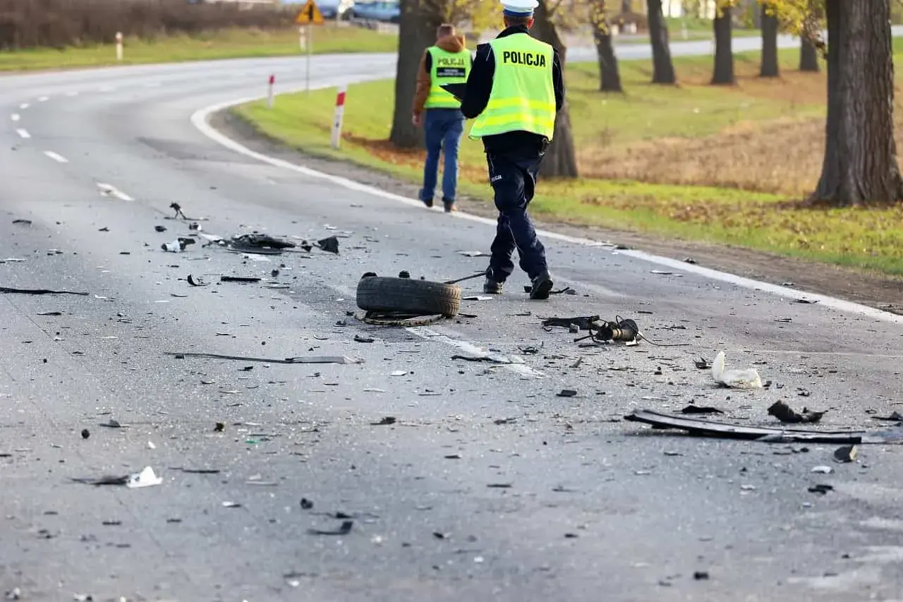 Na drodze po kolizji leżą szczątki, opona i elementy pojazdów. Policjanci w kamizelkach odblaskowych badają miejsce zdarzenia, być może szukając śladów ucieczki z miejsca kolizji.