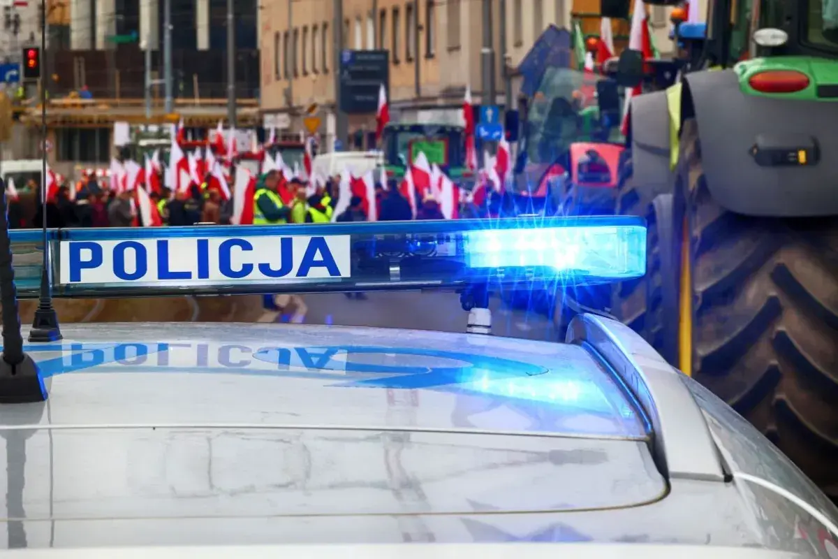 Policyjny radiowóz z włączonym sygnałem świetlnym na tle protestu rolników z flagami. Czy to jego jaka odprawa po 20 latach w policji?
