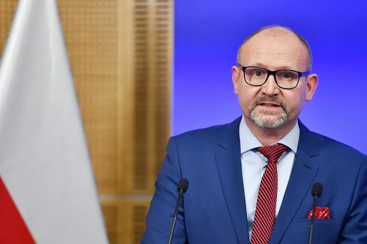 Prokurator krajowy w niebieskim garniturze, z okularami i czerwoną poszetką, przemawia przy mikrofonie. Obok widoczna flaga Polski.