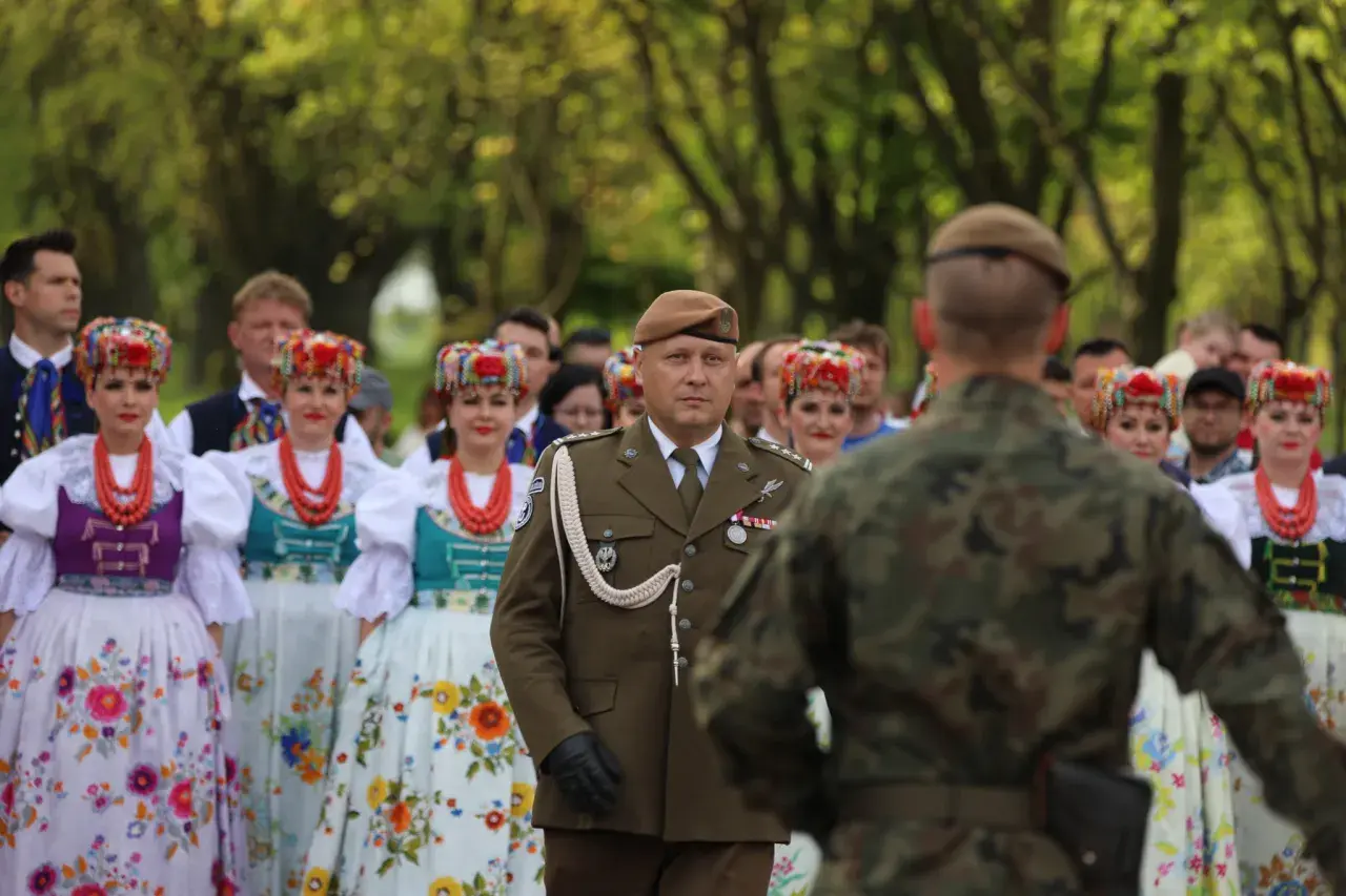 Żołnierz w brązowym capstrzyku i mundurze stoi przed grupą kobiet w tradycyjnych, kolorowych strojach ludowych.