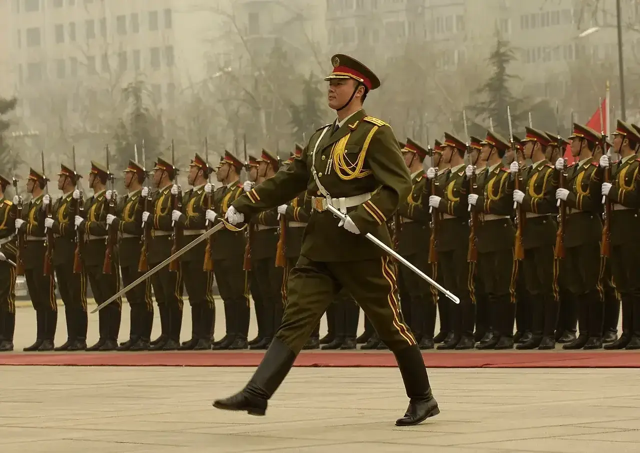 Kadet w galowym mundurze prowadzi defiladę, trzymając szablę. Za nim szeregi żołnierzy z karabinami.